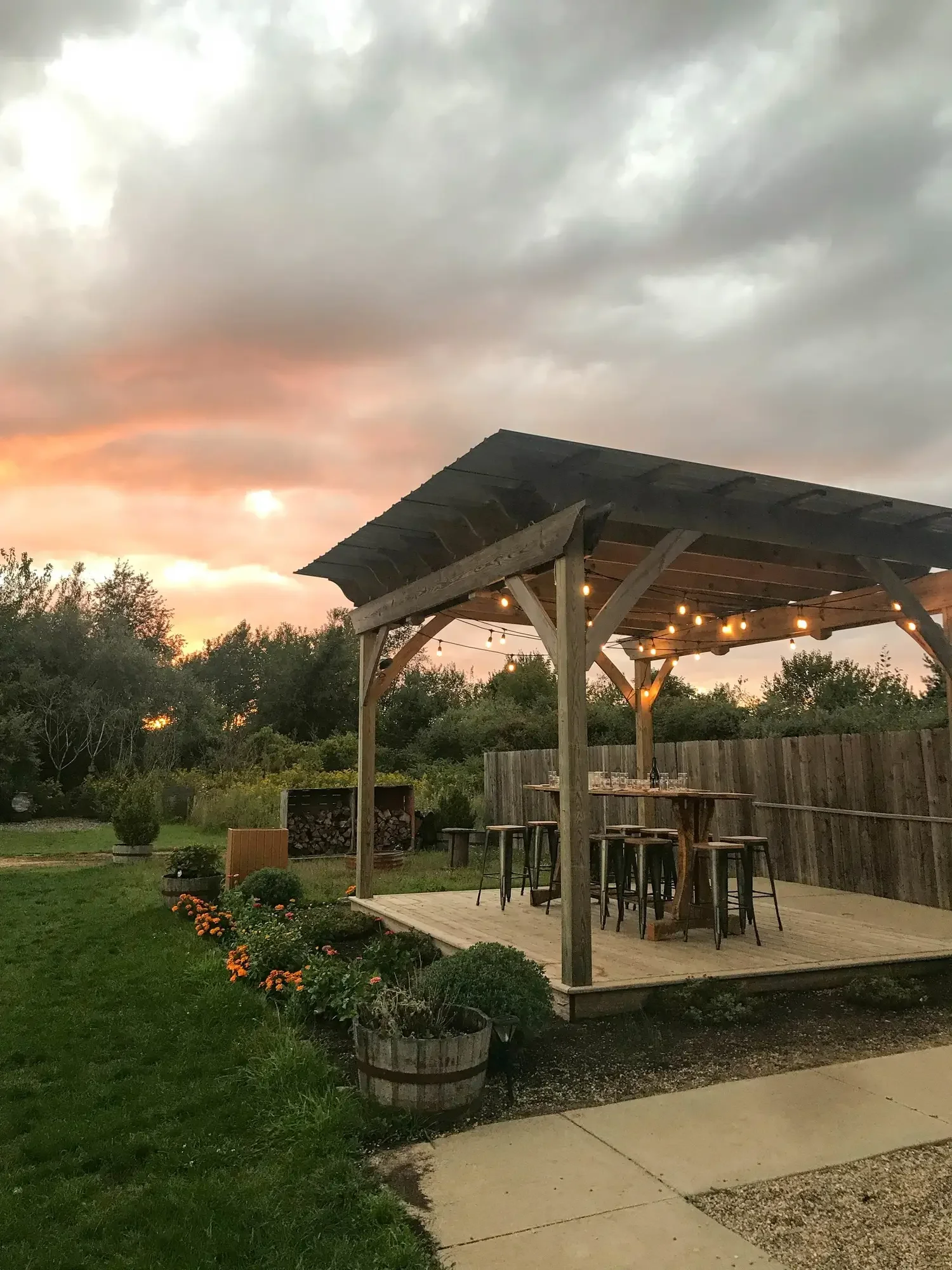 Outdoor patio with wooden pergola, string lights, a bar counter with stools, surrounded by greenery and flowers, under a cloudy sky at sunset.