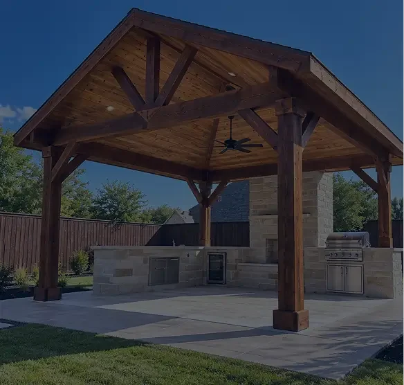 Outdoor kitchen with a wooden pavilion, built-in grill, stone countertops, and a ceiling fan, set on a concrete patio with a grassy yard and a wooden privacy fence.
