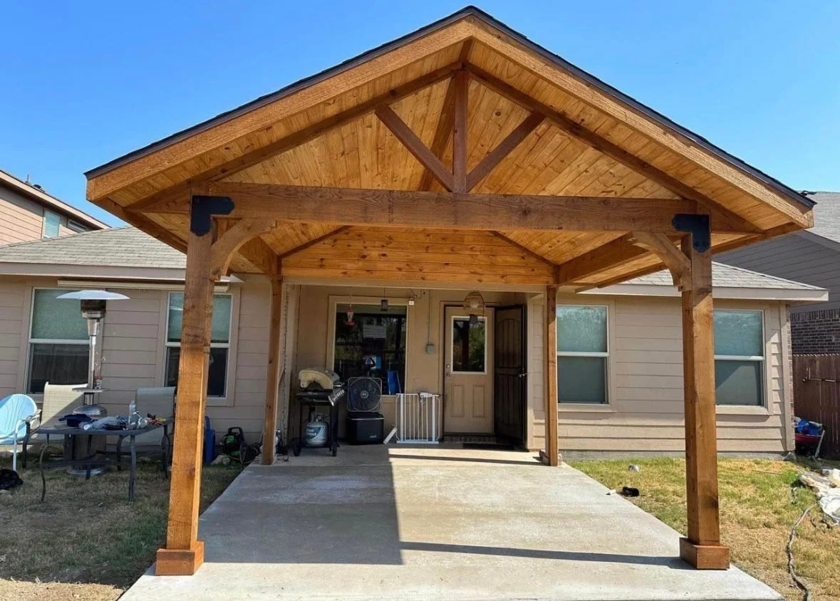 Wooden patio cover attached to the back of a house with concrete flooring underneath and outdoor furniture.