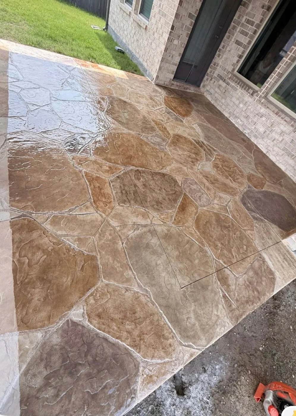 Newly installed decorative stone-patterned outdoor concrete patio next to a house with brick exterior walls and grass lawn, with construction tools visible.