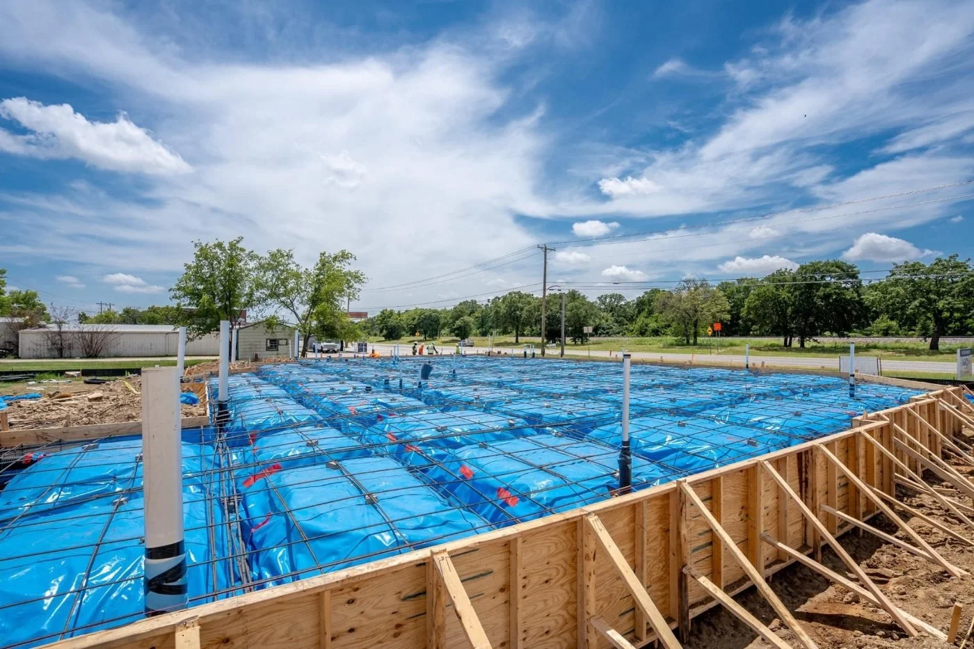 Construction site with wooden frame and blue waterproofing material, under a partly cloudy sky, with trees and power lines in the background.