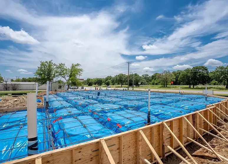 Construction site with a wooden frame foundation for a building, covered with blue plastic and metal rebar. In the background, there are green trees, a few buildings, and a partly cloudy sky.