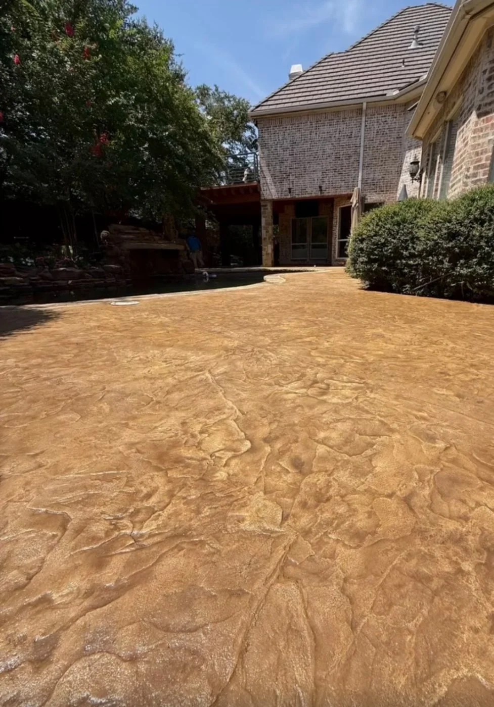 View of a backyard with a textured, stained concrete patio, some greenery including bushes and trees, and a two-story brick house with large windows and a patio door, under a clear blue sky.