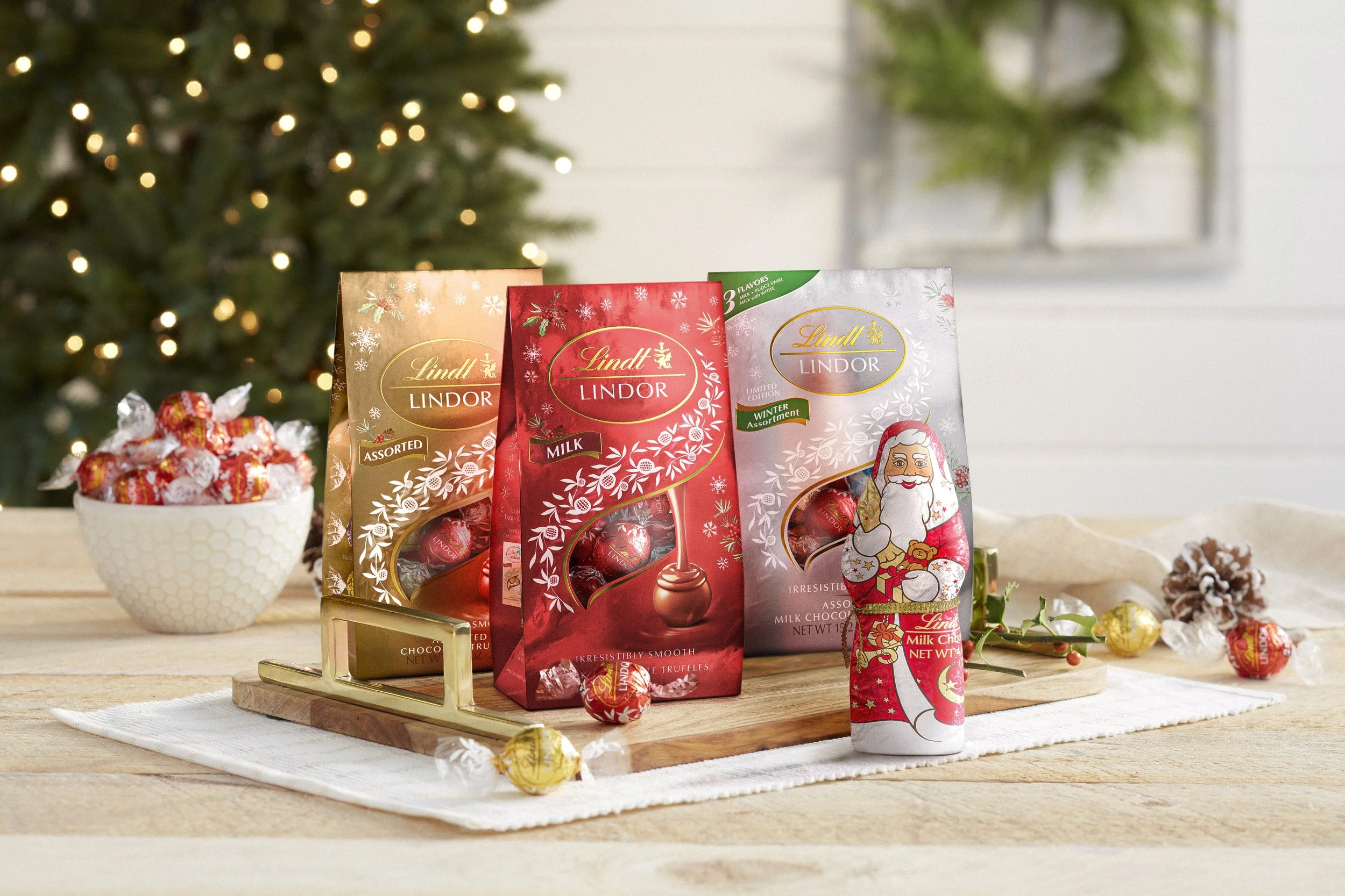 Assorted Lindt Lindor milk chocolates on a wooden tray with holiday-themed packaging, a bowl of wrapped candies, and Christmas decorations including a tree with lights.