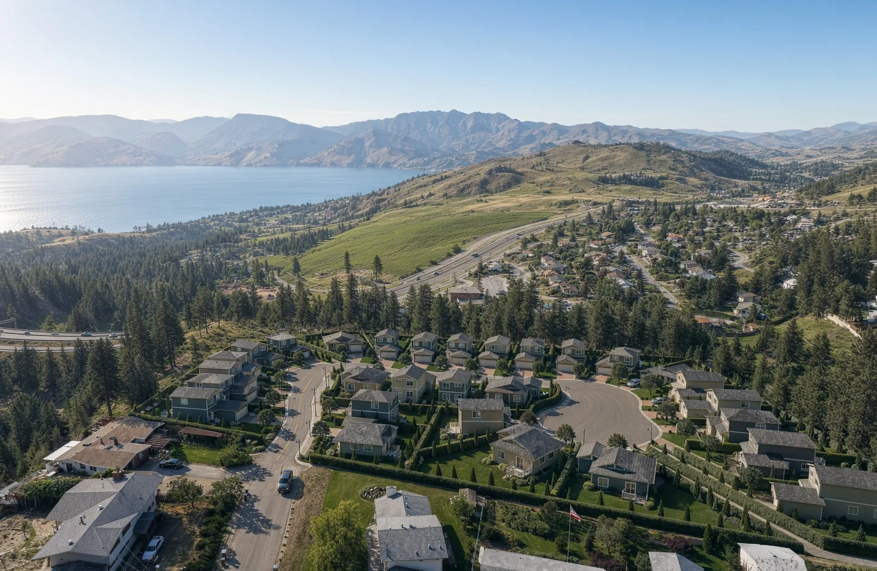 Aerial view of a lakeside suburban neighborhood in Kelowna, Canada with houses, trees, and winding streets, with mountains and a large body of water in the background.