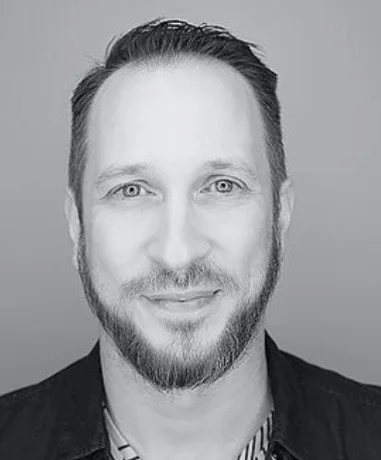 Black and white headshot of a man with short dark hair, facial hair, and a slight smile, wearing a dark jacket over a striped shirt.