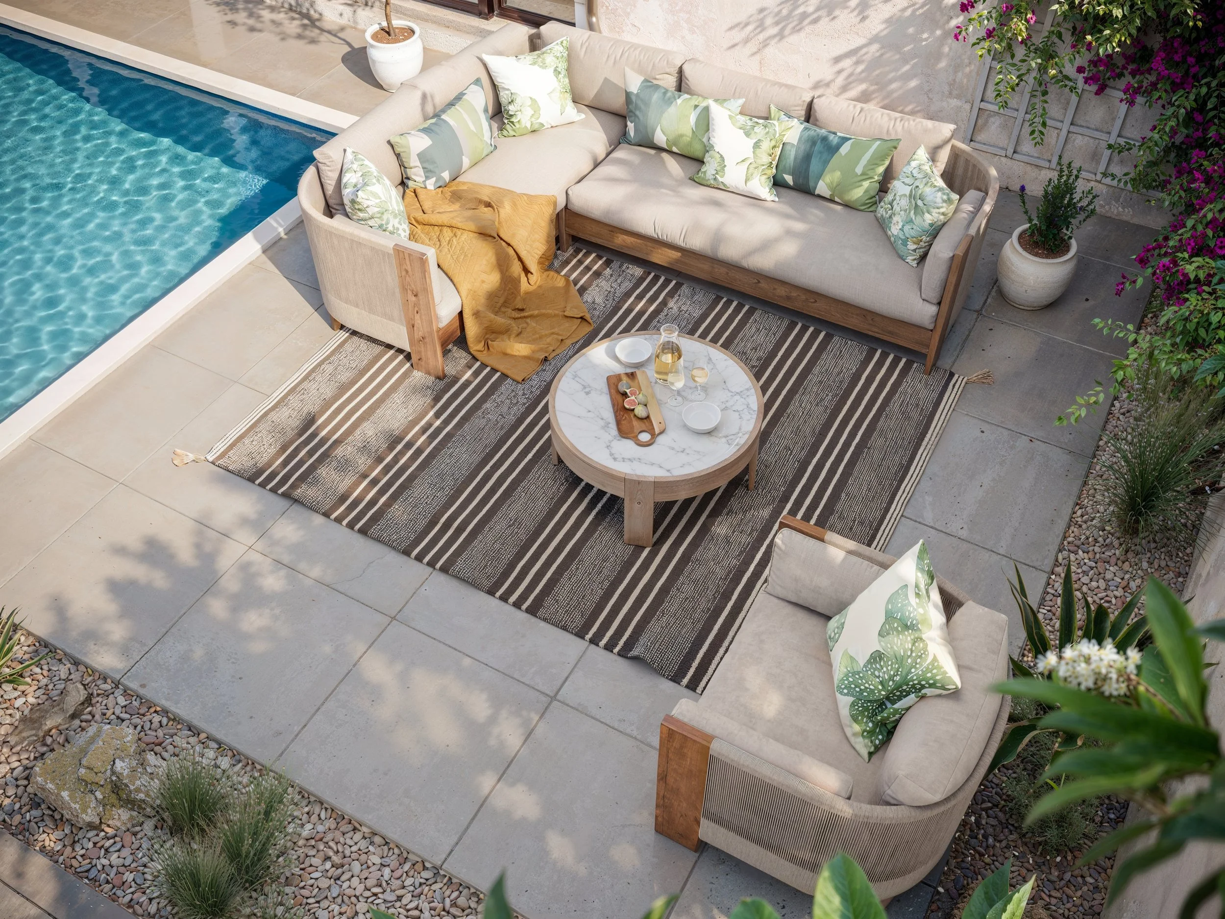 An outdoor seating area next to a swimming pool with a beige sectional sofa, decorative cushions with green and floral patterns, a round marble-topped coffee table with drinks and snacks, and against a wall with potted plants and greenery.