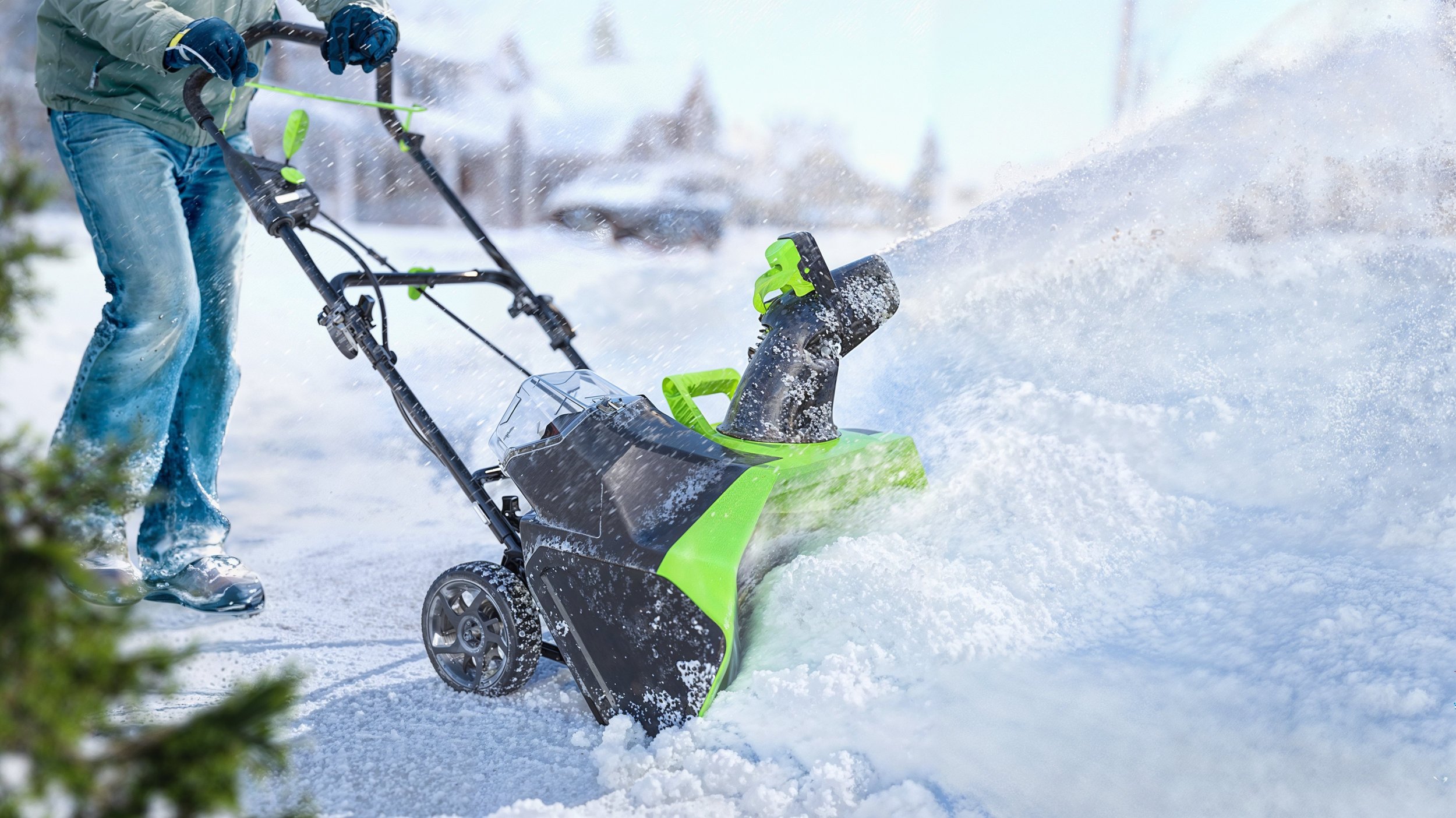 A person using a Greenworks snow blower to clear snow outside during winter, with snow flying around.