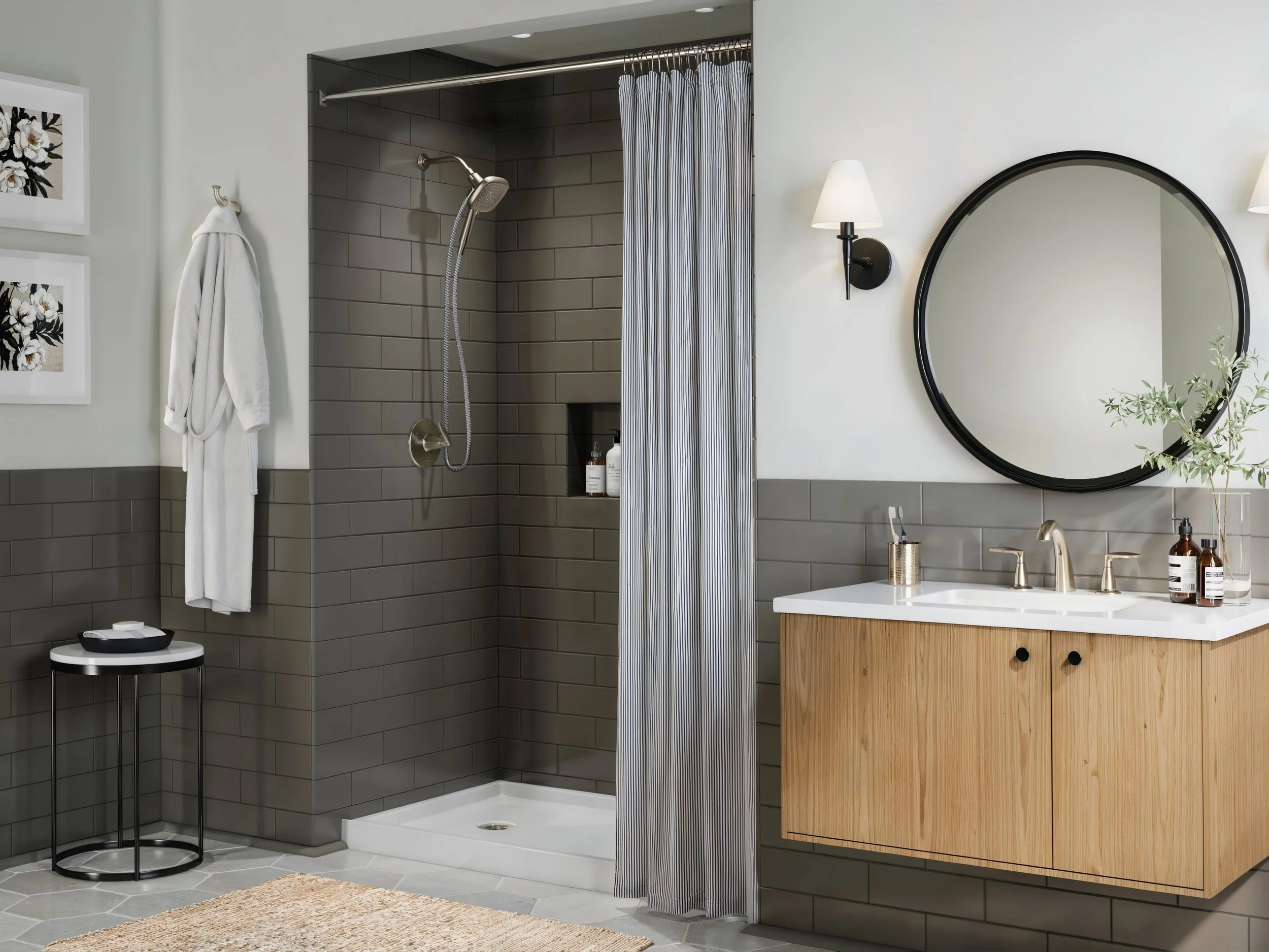 Modern bathroom with a walk-in shower featuring gray tile walls and a striped shower curtain. A white bathrobe hangs on a hook, and a small circular table with a black and white design is nearby. There is a large round mirror above a wooden vanity with a white countertop, and black wall-mounted lamps flank the mirror. Decor items and toiletries are placed on the vanity.