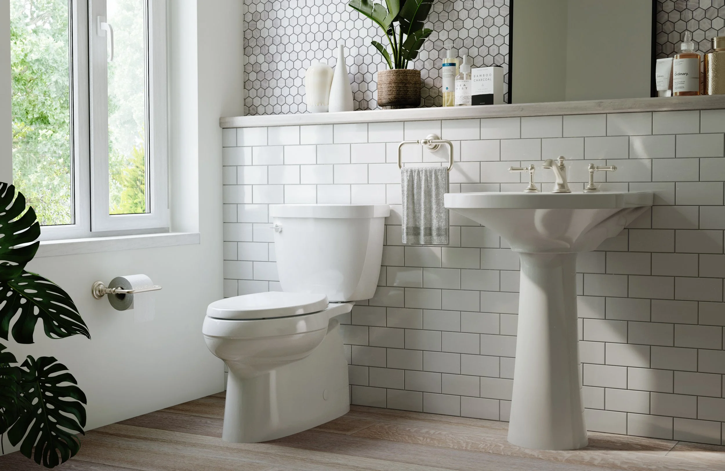 A modern bathroom featuring a white toilet, white subway-tile walls, a white pedestal sink, and a large window with a view of greenery outside. Decor includes a potted plant, vases, and personal care products on a marble ledge.