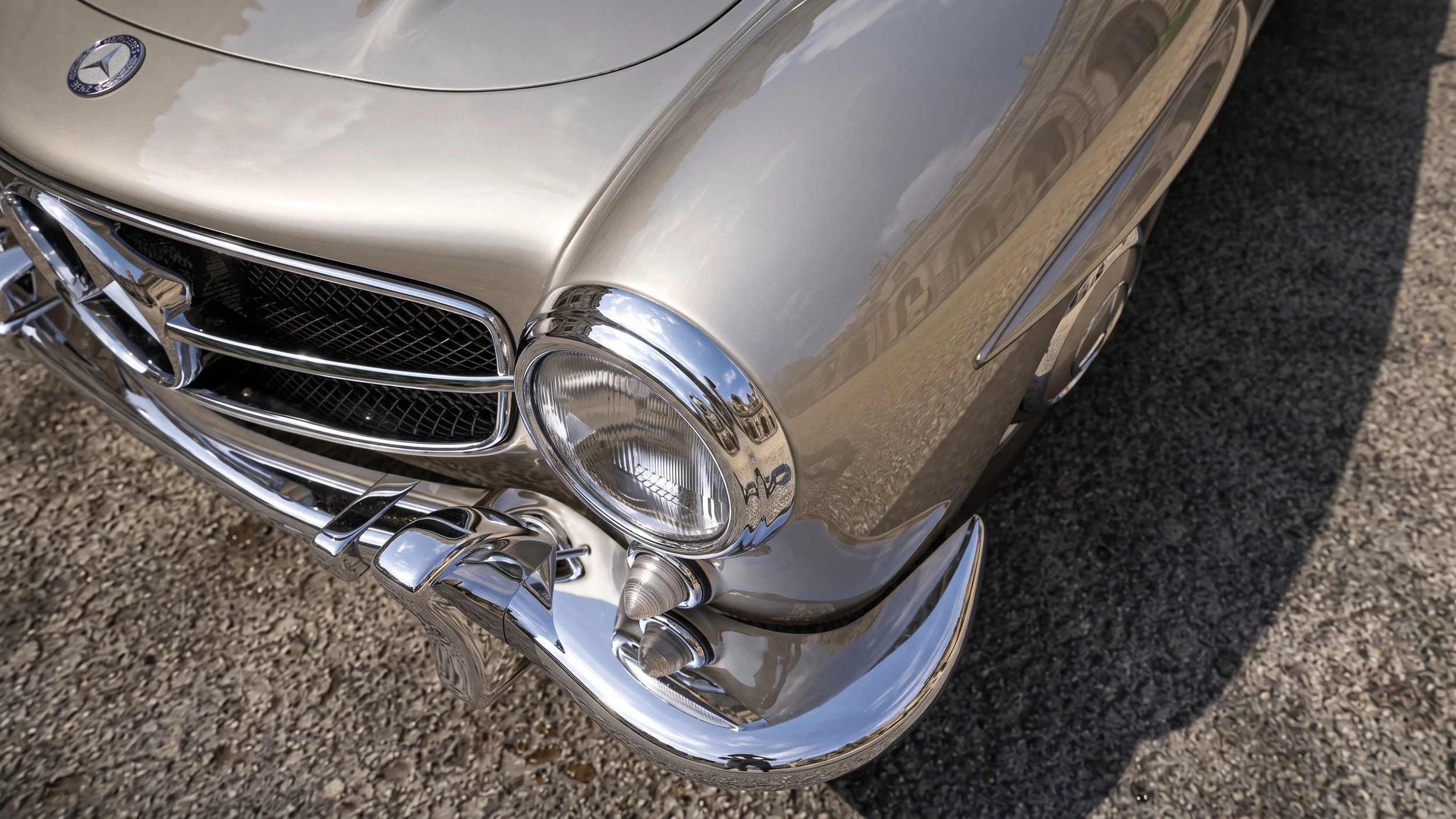 Close-up image of a vintage silver Mercedes-Benz car front, showing chrome details, headlight, and the Mercedes emblem on the hood, parked on a gravel surface.