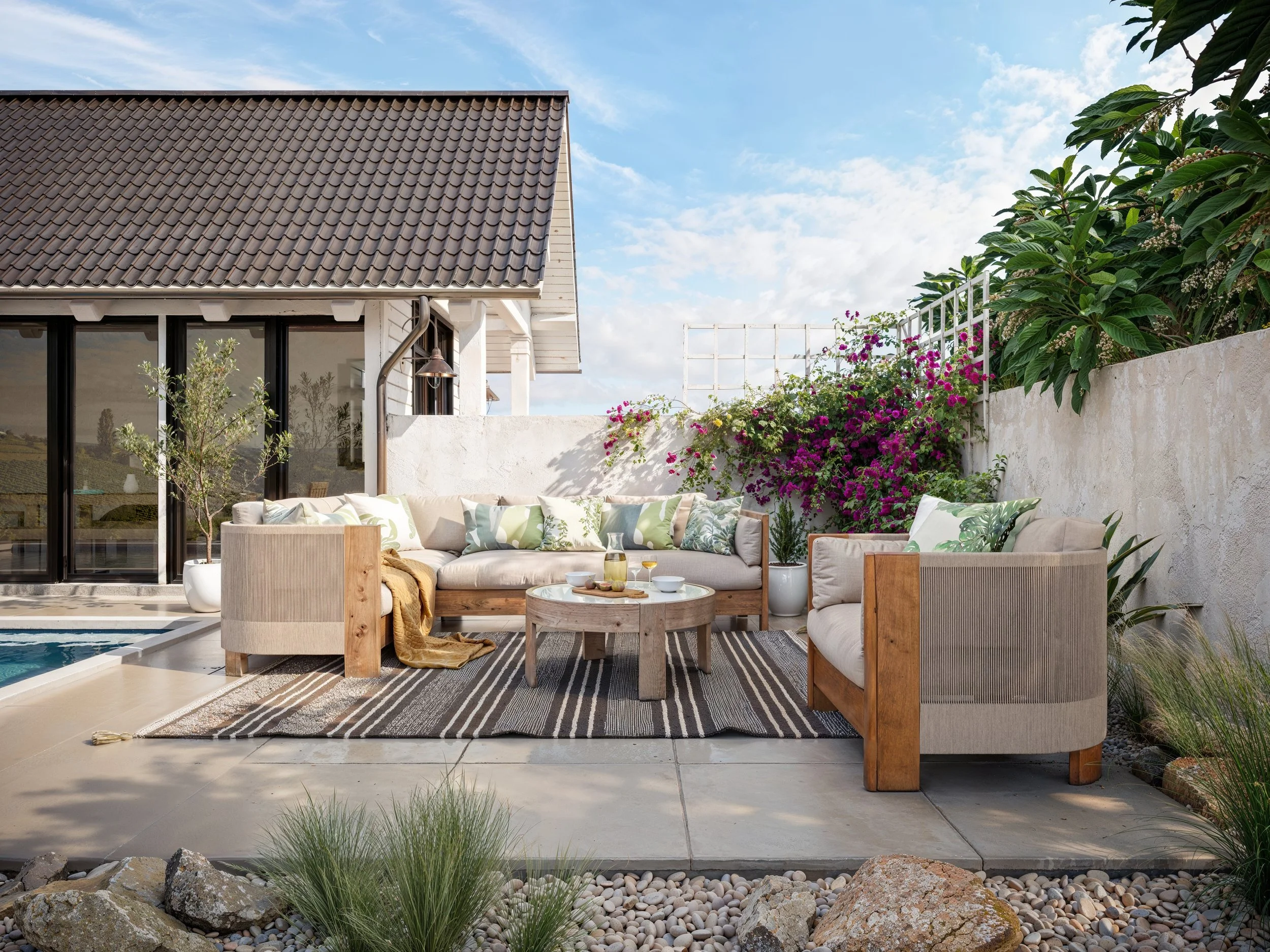 A cozy outdoor West Elm patio furniture, green and patterned pillows, a bamboo side table with drinks, a striped rug, and lush plants and flowers in a white-walled garden, with a house with a tiled roof in the background and a partly cloudy sky.
