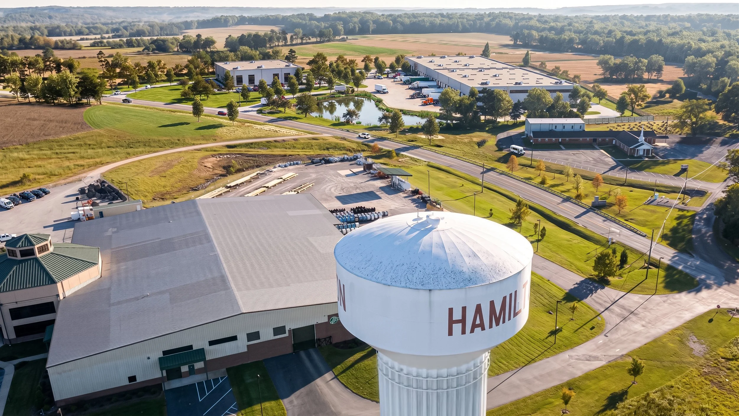 Aerial view of an industrial complex with a water tower labeled 'HAMIL' in front, surrounded by parking lots, a pond, and green fields with trees.