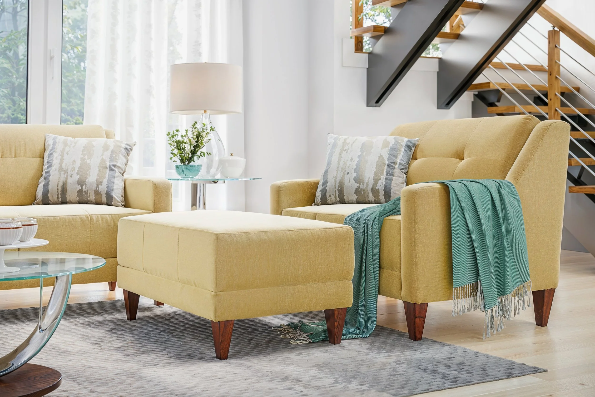 Living room with yellow-colored upholstered Laz-y-Boy sofa and matching ottoman, throw pillows, a glass table with a vase of white flowers, and a teal throw blanket. Large windows with white curtains