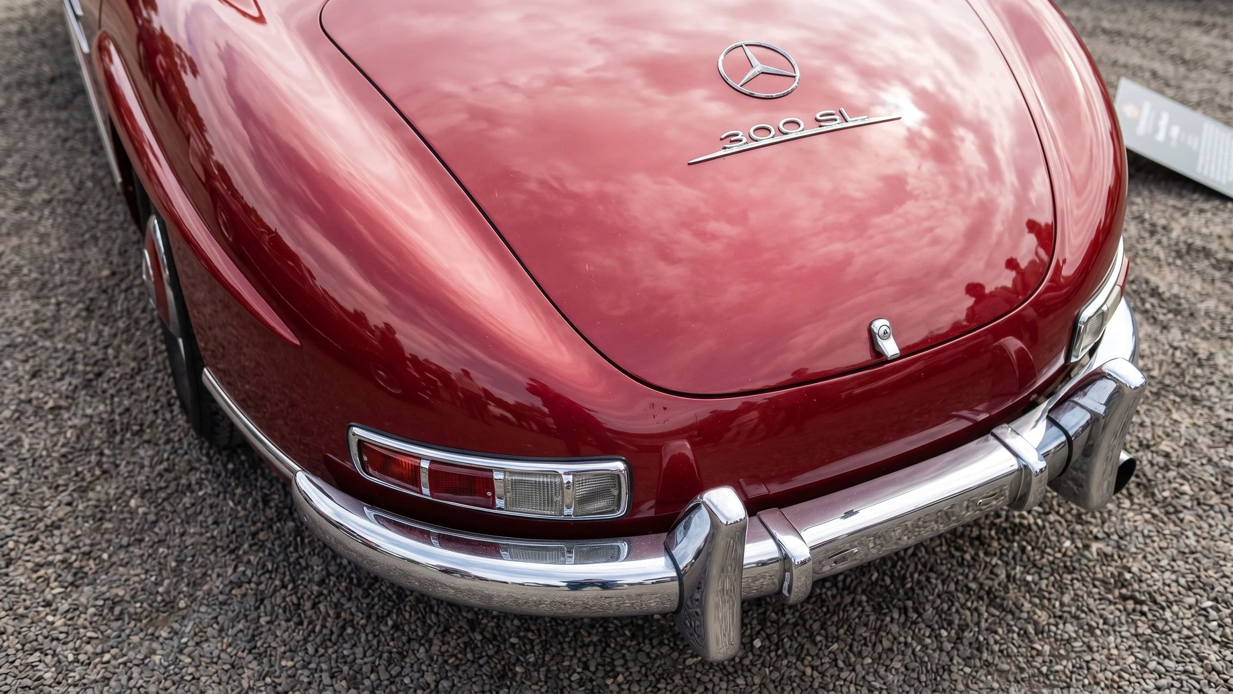 Red vintage Mercedes-Benz 300 SL car with chrome bumper and emblem, parked on gravel surface.