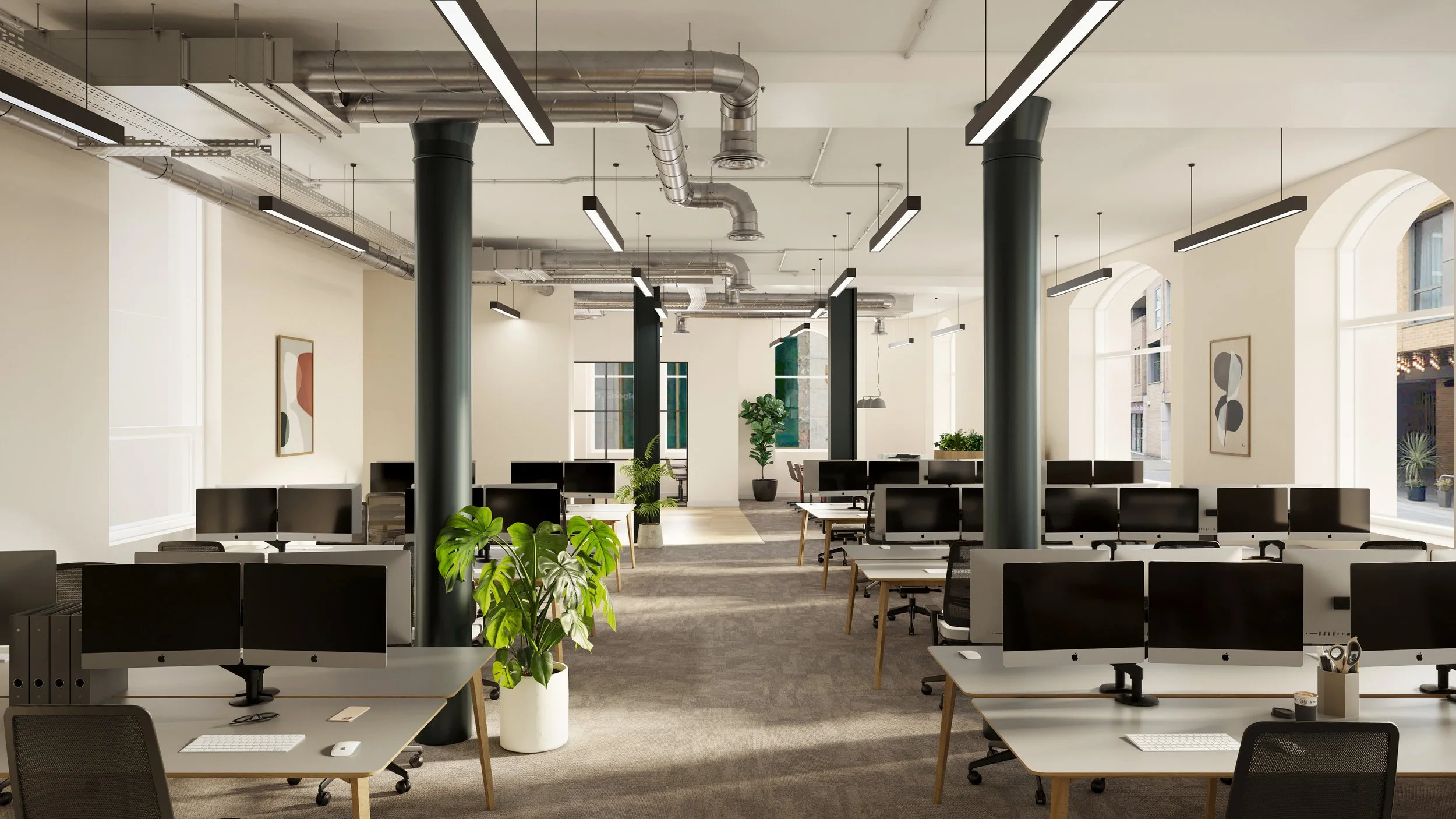 An open-plan modern office space with rows of desks featuring desktop computers, some with dual monitors, and office chairs. The room has large windows letting in natural light, decorated with artwork on the walls and several green potted plants. The