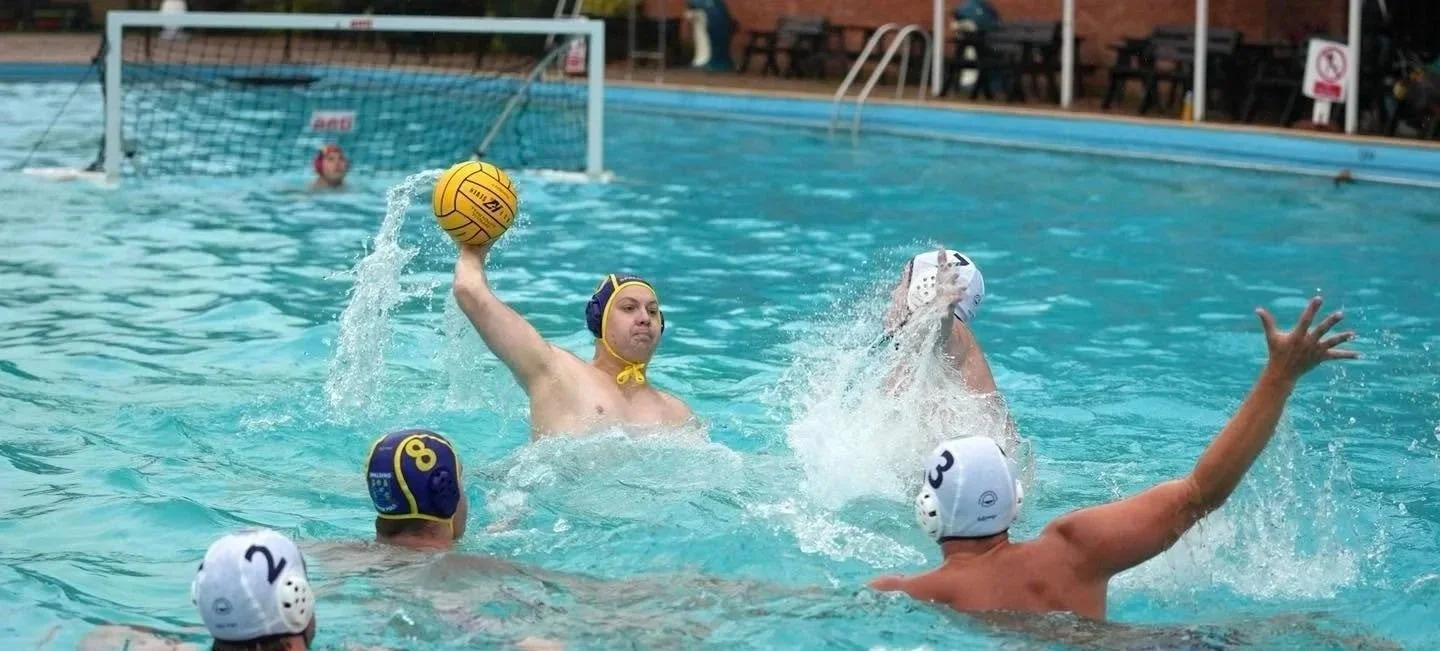 Water polo players competing in a game, with one player in the act of throwing the ball towards the goal, while others defend or position themselves in the pool.