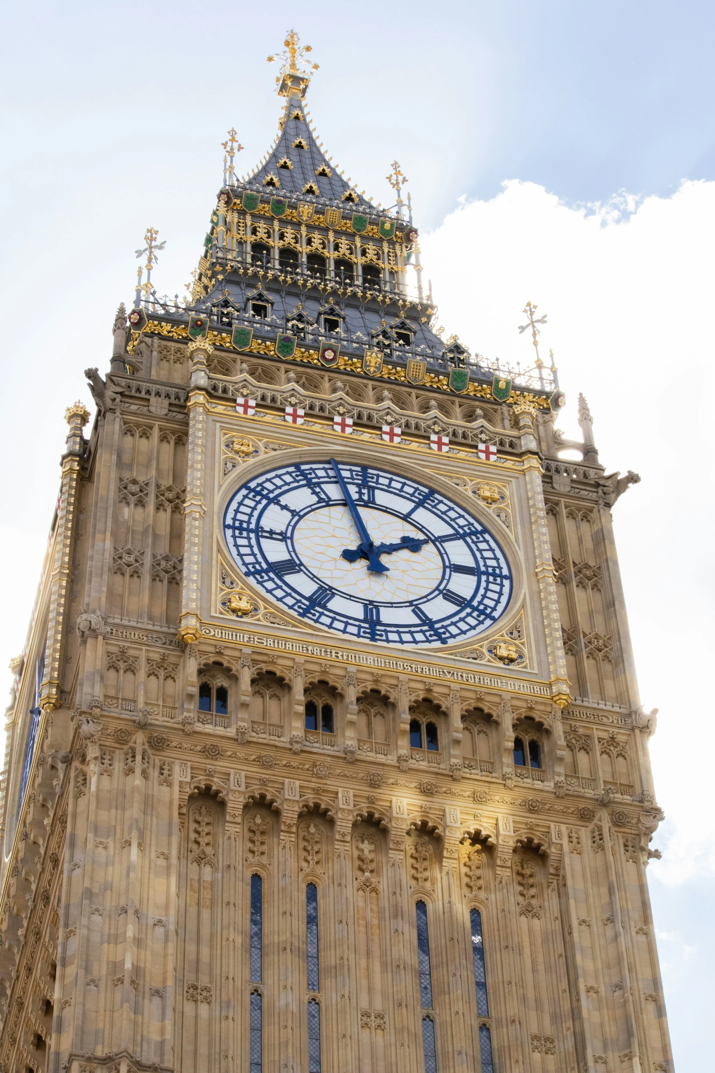 Close-up of the Queen Elisabeth II clock tower showing Big Ben of the Palace of Westminster in London, showing the ornate Gothic architecture and the clock face with the time showing approximately 1:55 PM. The sky is partly cloudy in the background.