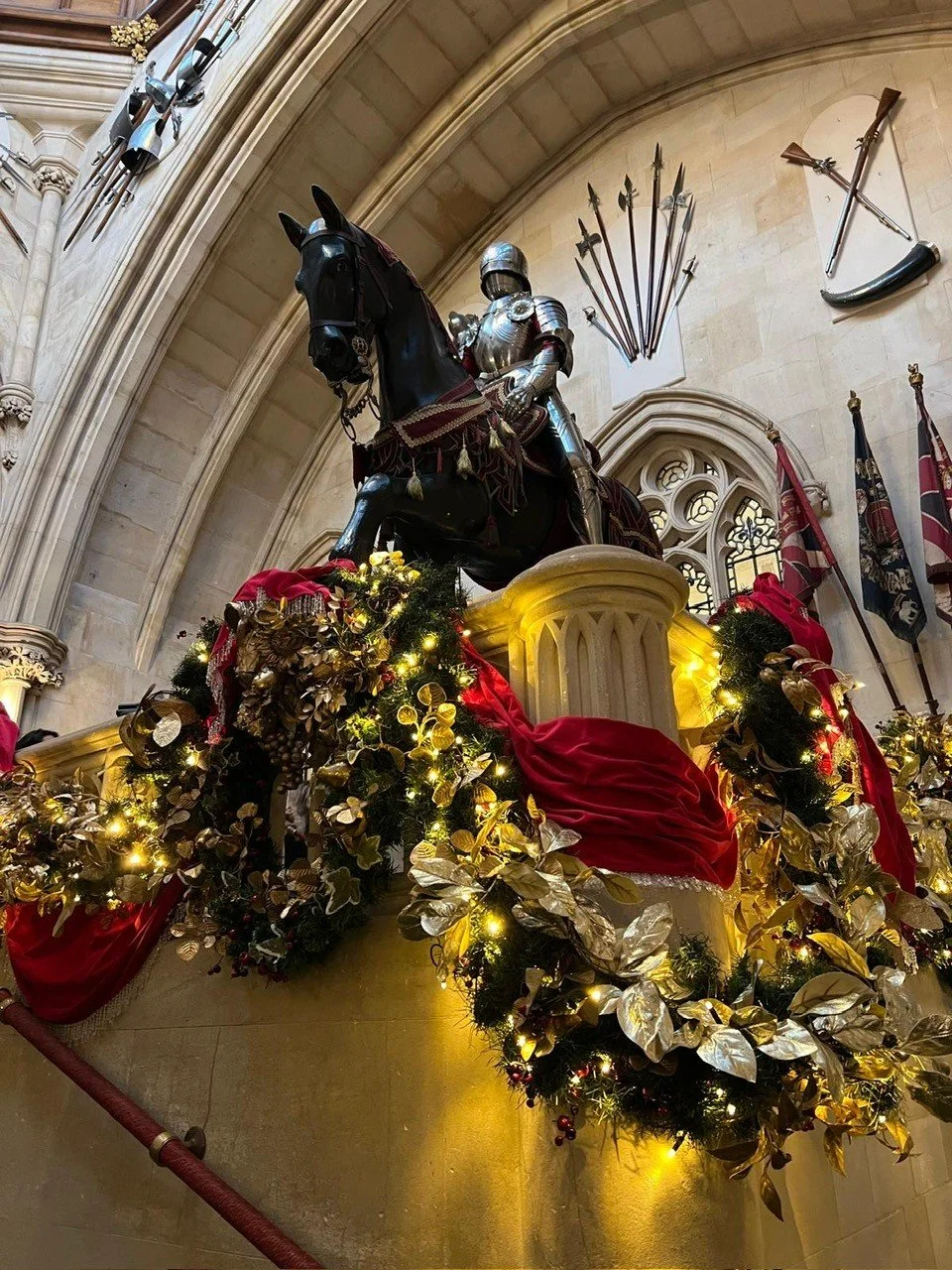 Decorated Christmas display with a black knight on horseback, Christmas garland, gold leaves, lights, and red fabric, inside a Gothic-style hall with medieval weapons and shields on the wall.