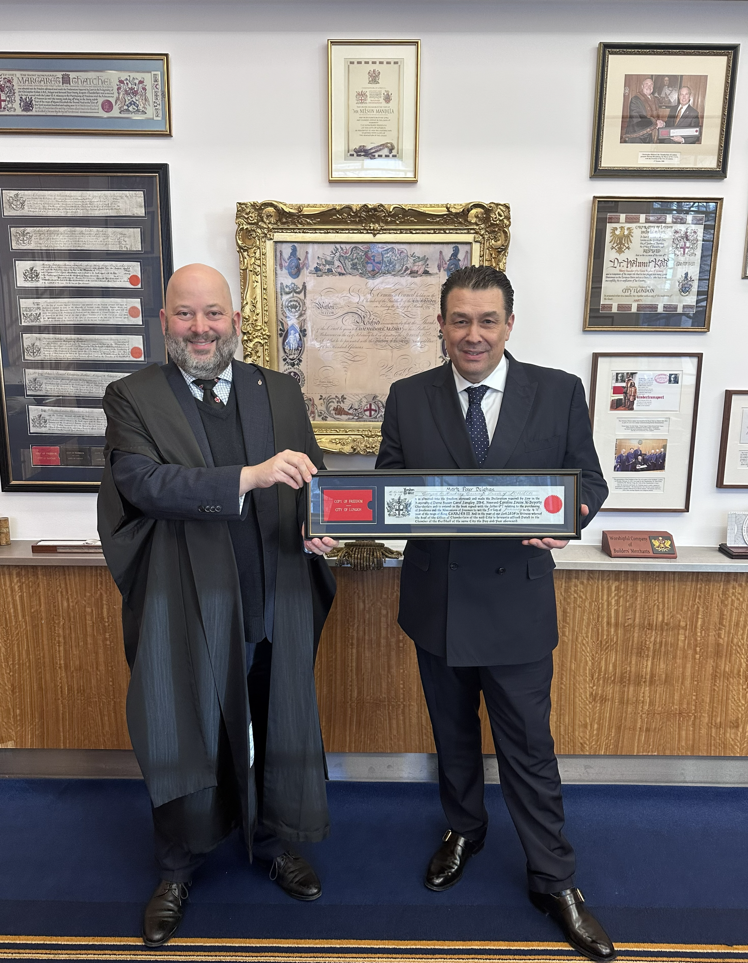 Two men in suits standing in front of a wall with framed certificates and photos, holding a framed certificate together, smiling at the camera.