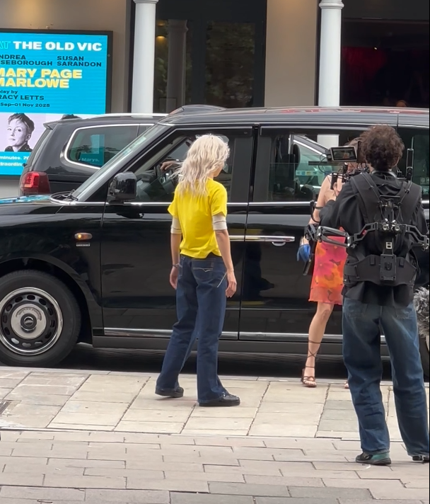 Two women stand near a black SUV on a city sidewalk. One woman with blonde hair in a yellow shirt and blue jeans faces the vehicle, while the other woman, with darker hair, wears a black top and colorful skirt and holds a water bottle. A cameraman with a vide camera records the scene. In the background, a digital billboard displays information about a presentation featuring Mary Page Marlowe.