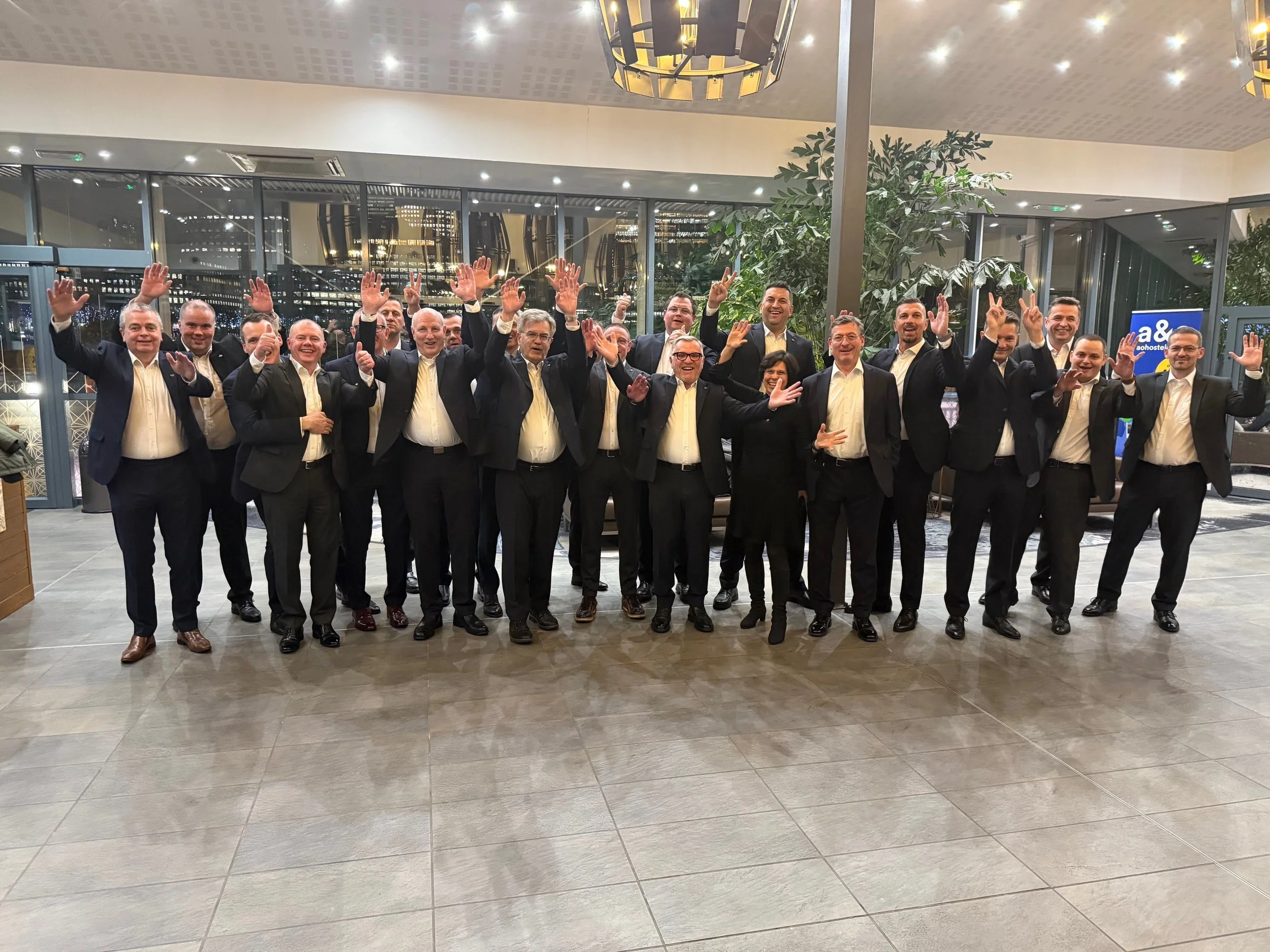 Group of approximately 16 people in formal attire standing inside a building with large windows, decorations, and city lights visible outside, celebrating and smiling with some raising their hands.
