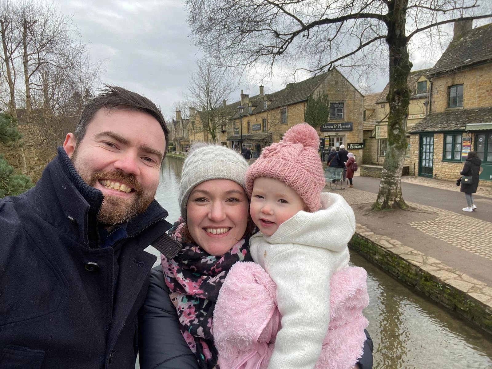A smiling family of three taking a selfie outdoors near a canal in a quaint town with stone buildings, trees, and pedestrians in the background, all dressed warmly for cold weather.