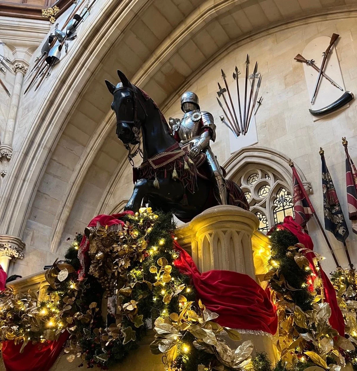 A statue of a knight in armor on a horse, decorated with red fabric and gold accents, surrounded by Christmas decorations including gold and red foliage, with swords and flags mounted on the wall behind, inside a grand stone building.