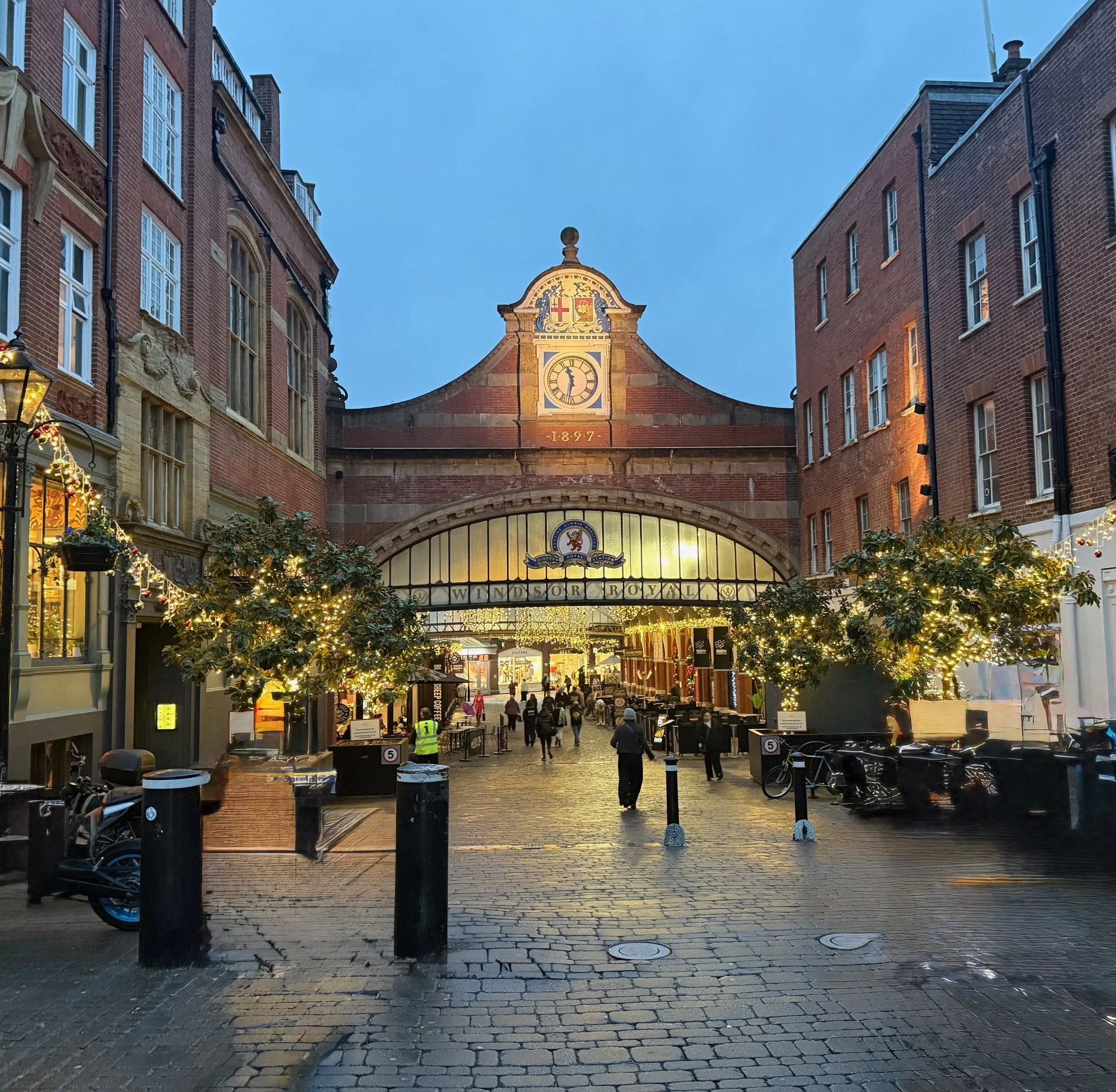 Windsor Royal Station entrance decorated with festive lights and greenery, with people walking along cobblestone street at dusk, historic buildings on either side, and a blue sky overhead.