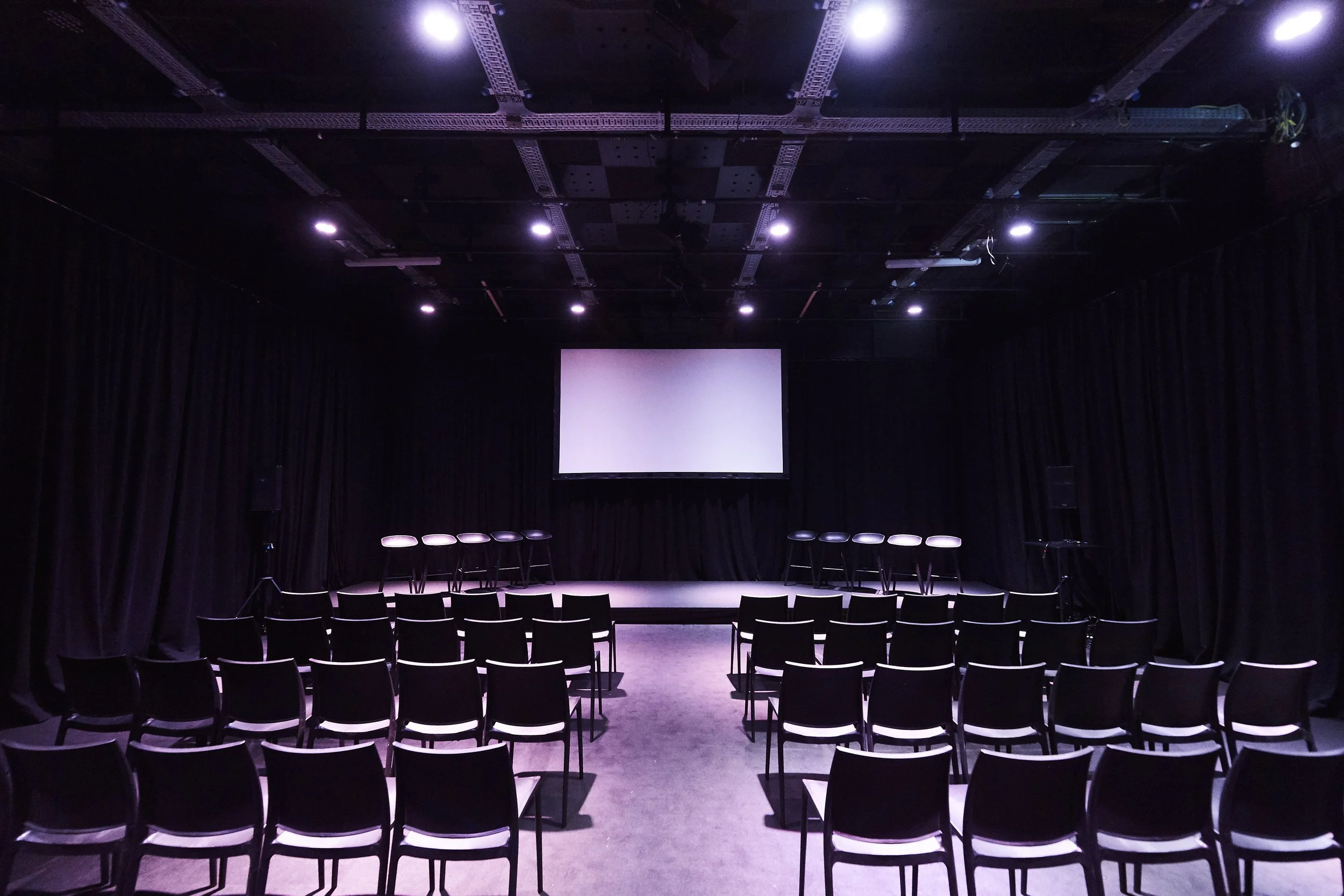 Empty auditorium or theater with rows of chairs facing a large screen or stage, dim lighting, and black curtains on sides.