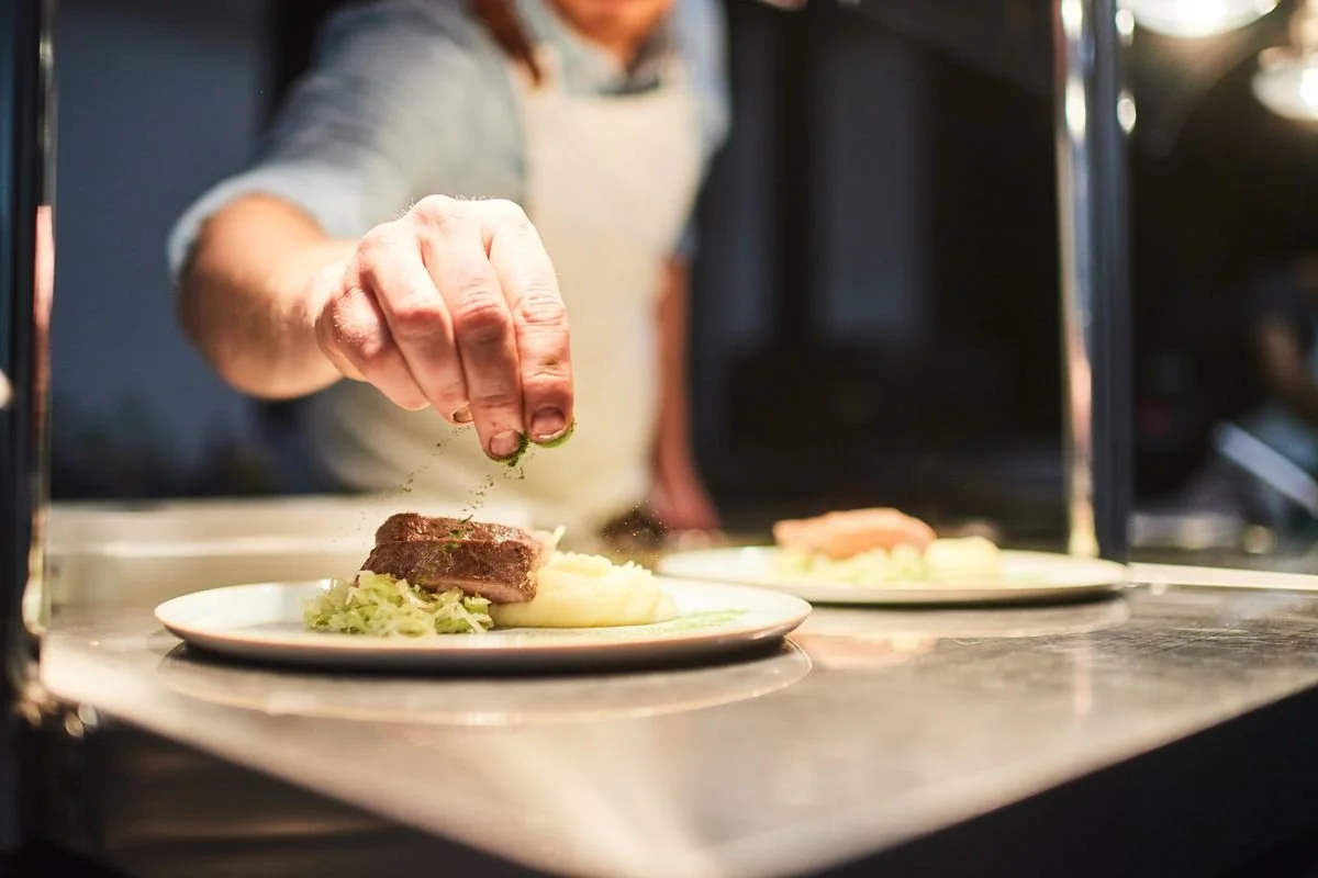 A person sprinkling herbs or seasoning on a plated serving of food that includes mashed potatoes, a cut of steak, and vegetables.