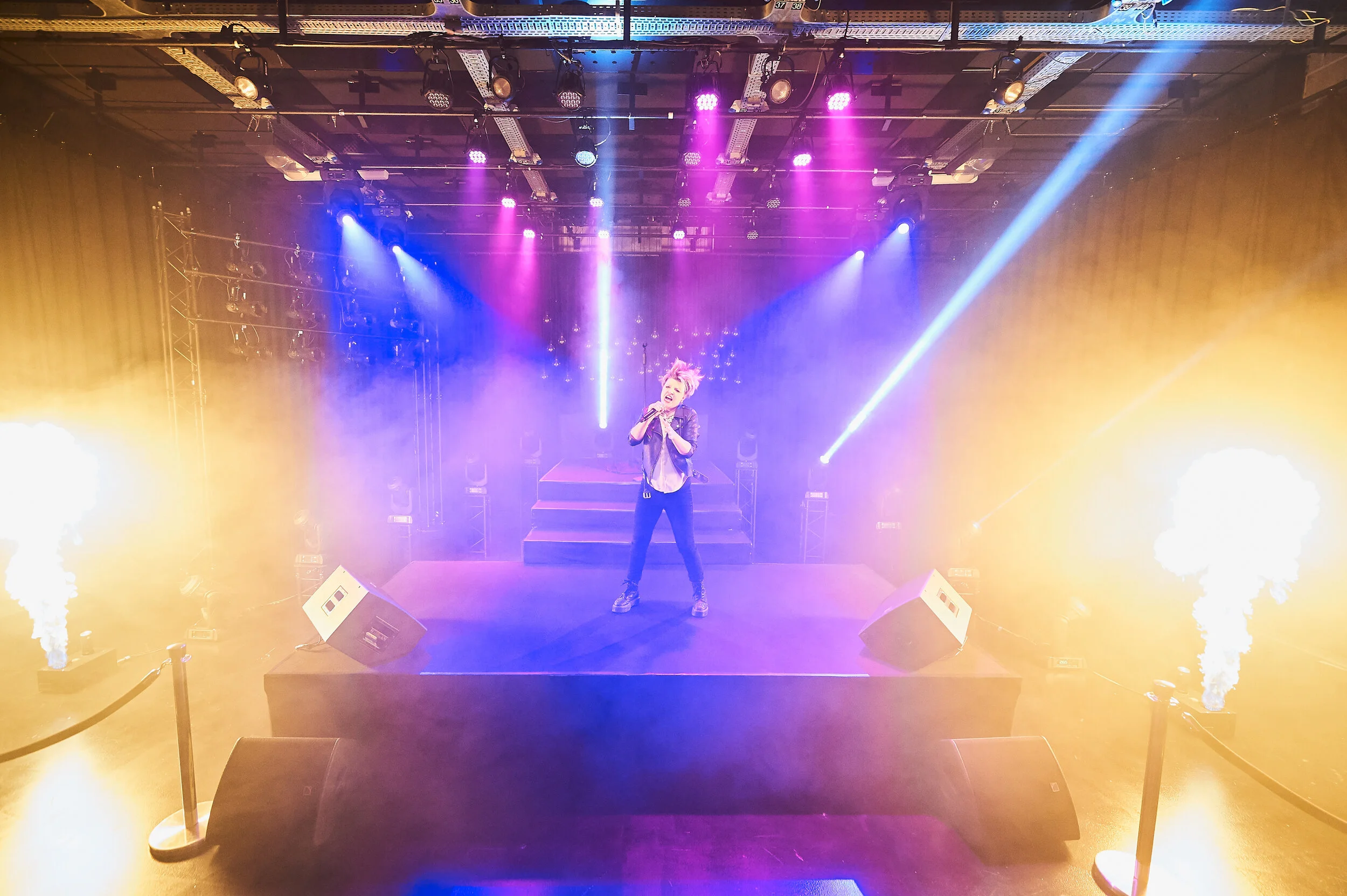 Performer singing on stage with colorful lights, fog, and pyrotechnics in a concert setting.