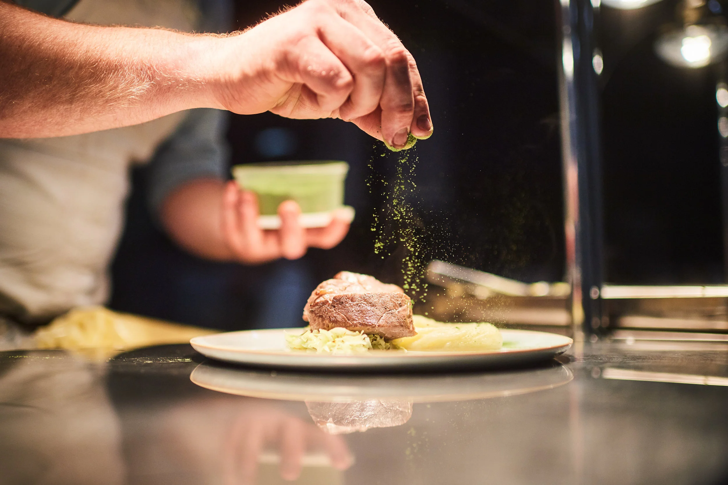 Chef seasoning a plated dish with green powder, with a person holding a container in the background.
