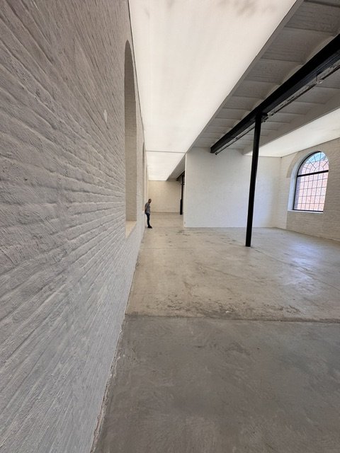 An empty industrial-style interior with high ceilings, exposed beams, large arched window, and minimal decor.