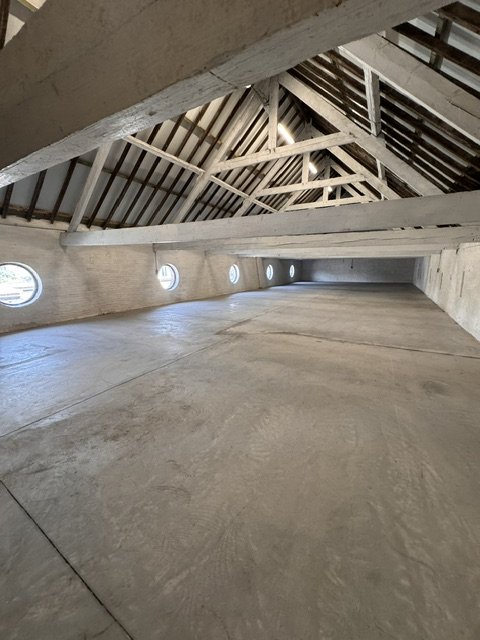 Empty attic space with small round windows along the wall, exposed wooden roof trusses, and concrete floor.