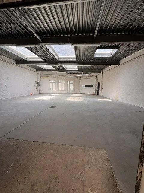 Empty industrial or commercial space with high ceiling, exposed metal roof, concrete floor, and large windows at the back.