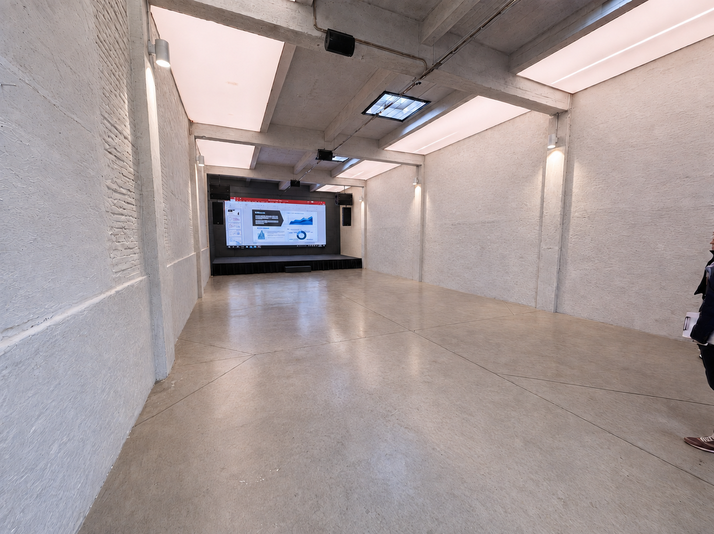 Empty room with white textured walls, beige floor, ceiling lights, and a large screen displaying charts and graphs at the far end.
