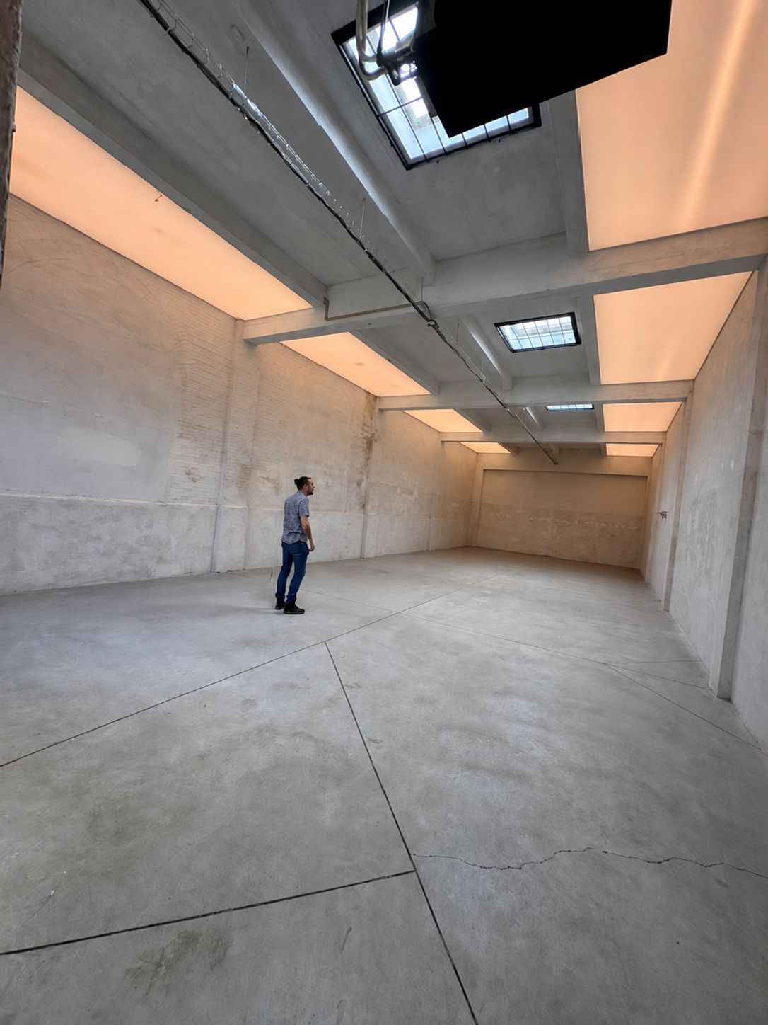 A man standing in an empty, industrial-style room with concrete walls and floors, illuminated by skylights on the ceiling.
