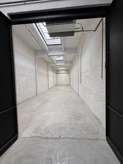 View of an empty, narrow industrial corridor with concrete walls and floor, overhead skylights, exposed piping, and a metal door frame in the foreground.