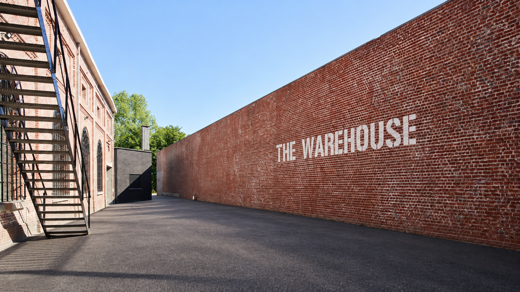 View of a brick warehouse with a large painted sign reading 'The Warehouse' on the side, an outdoor fire escape on the left, and a paved area in front under a clear blue sky.