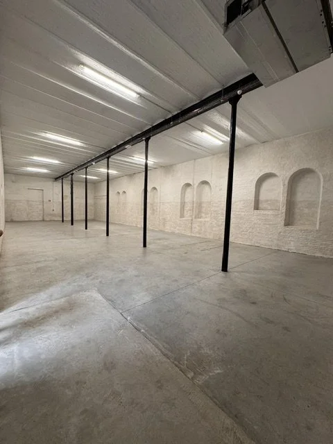 Empty industrial warehouse with concrete floors, white brick walls with arched windows, and black support columns.