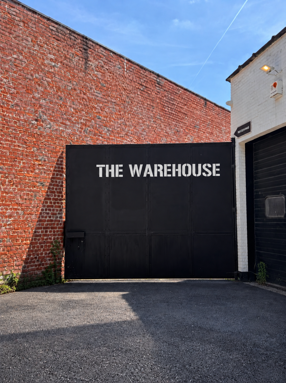 Black gate with white text that reads 'THE WAREHOUSE,' set between red brick and white walls, with a clear blue sky overhead.