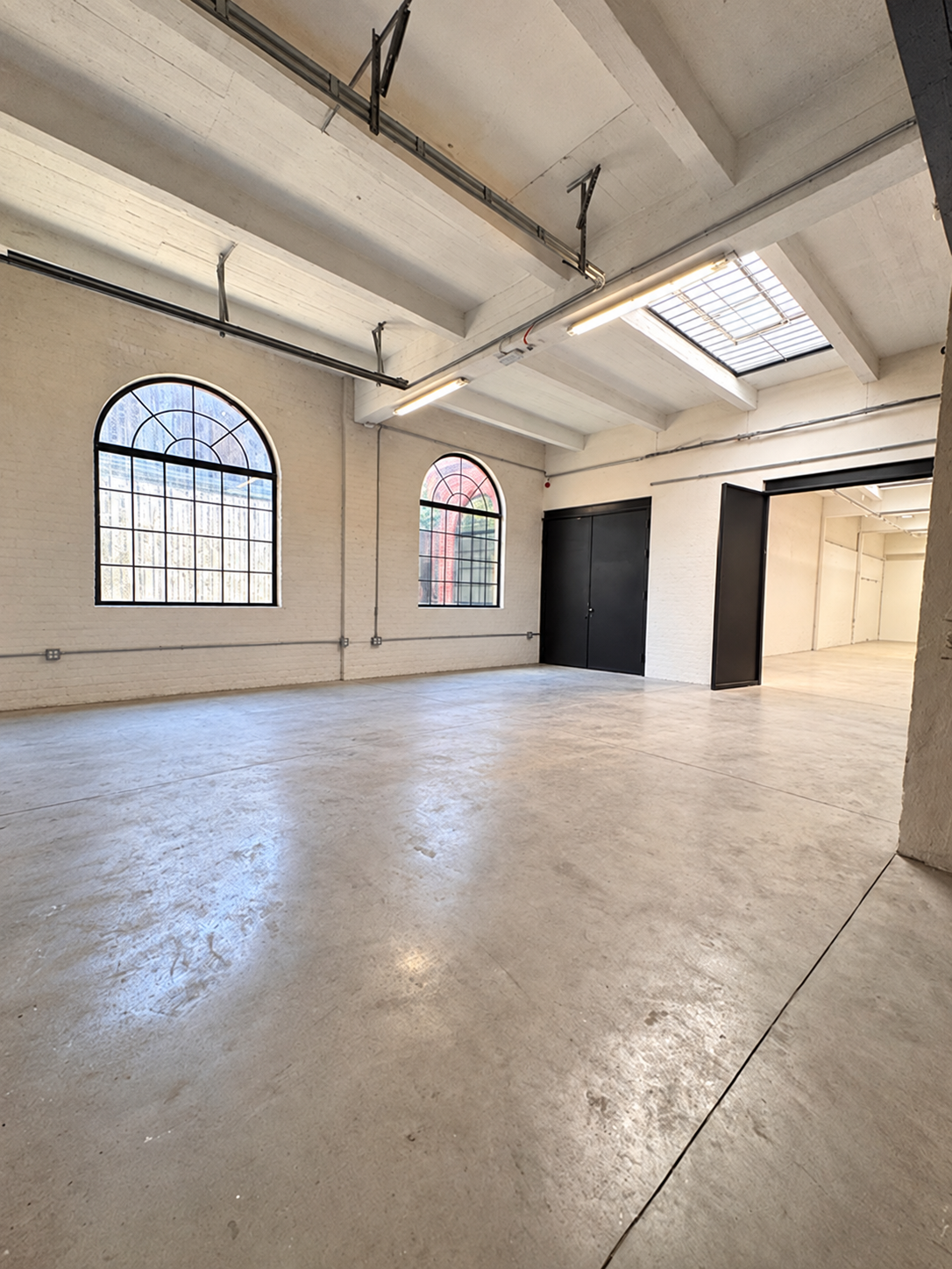 Open, spacious industrial-style room with white brick walls, large arched windows, concrete floors, and exposed overhead piping and lighting.