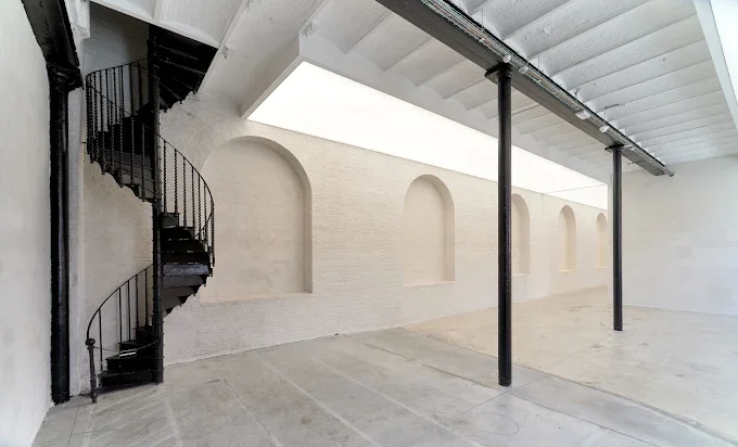Open industrial-style outdoor area with a black spiral staircase on the left, white brick walls with three arched recesses, a concrete floor, and black metal supports holding up a ceiling or overhang.