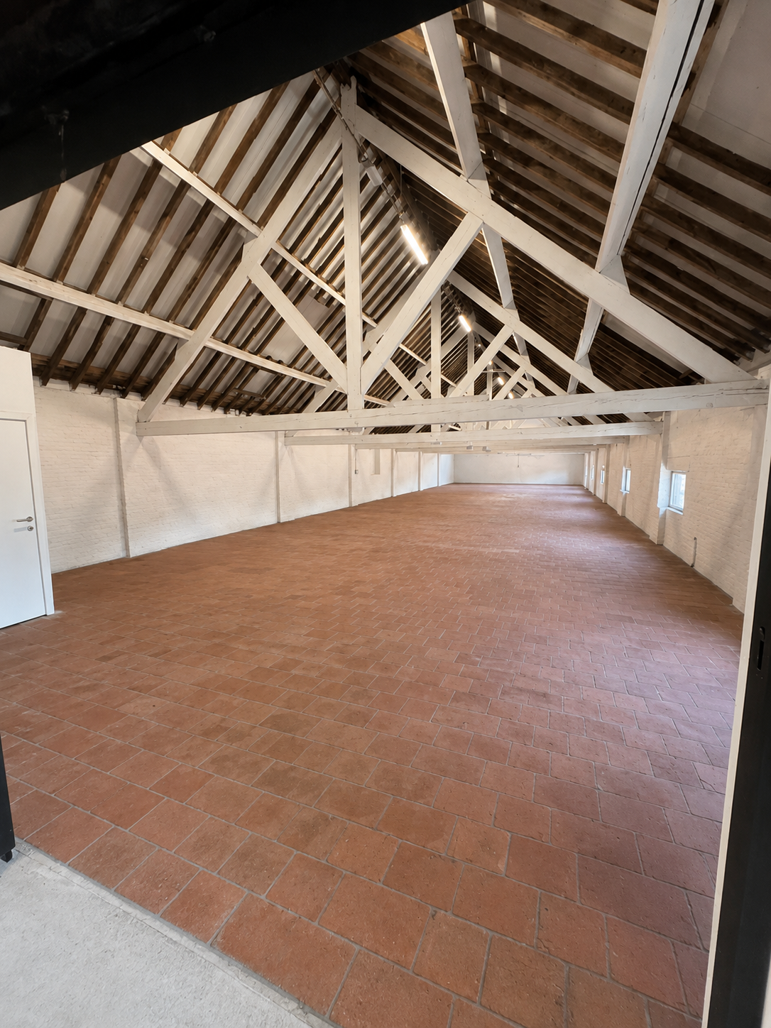 Empty attic space with exposed wooden beams and terracotta tile flooring, white brick walls, and small windows.