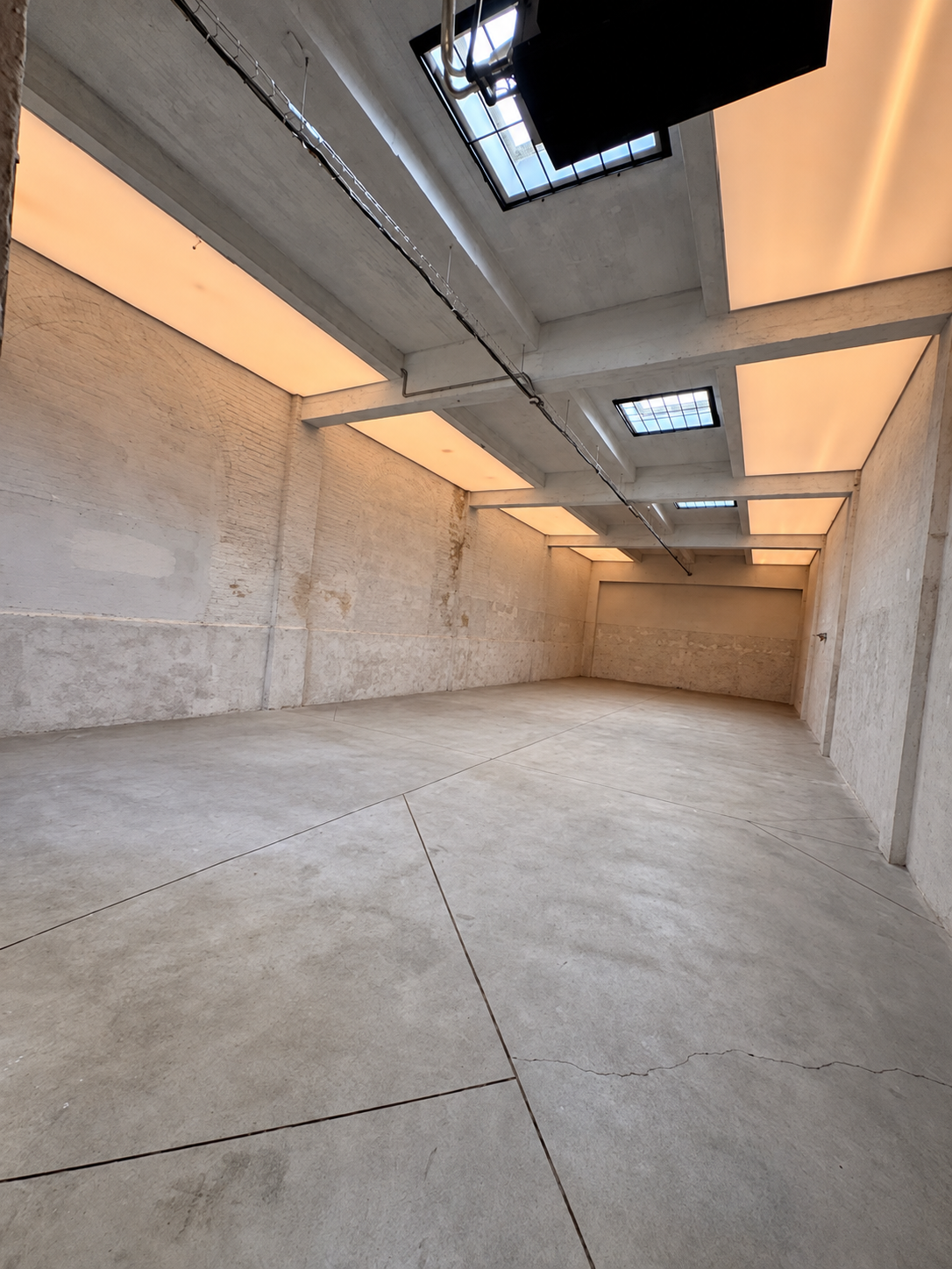 Empty industrial-style room with concrete walls and floor, large skylights, and warm ceiling lighting.