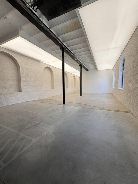 Empty room with curved brick walls, large arched windows, and a sloped ceiling with skylights, supported by black pillars.
