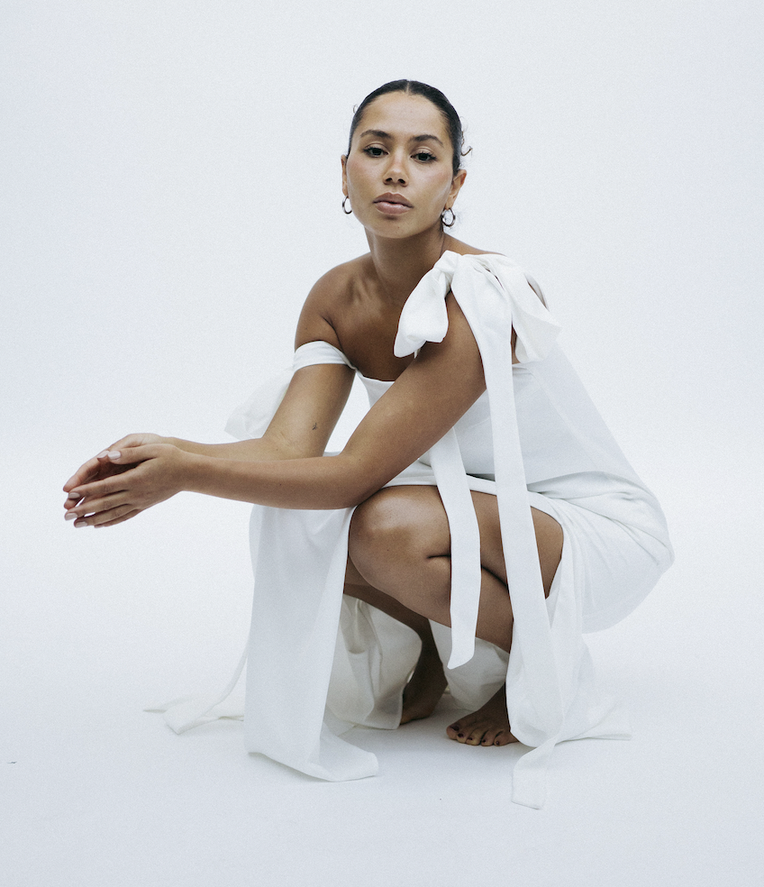 A woman with dark hair in a bun wearing a white dress with bow and shoulder details, squatting on a white background.