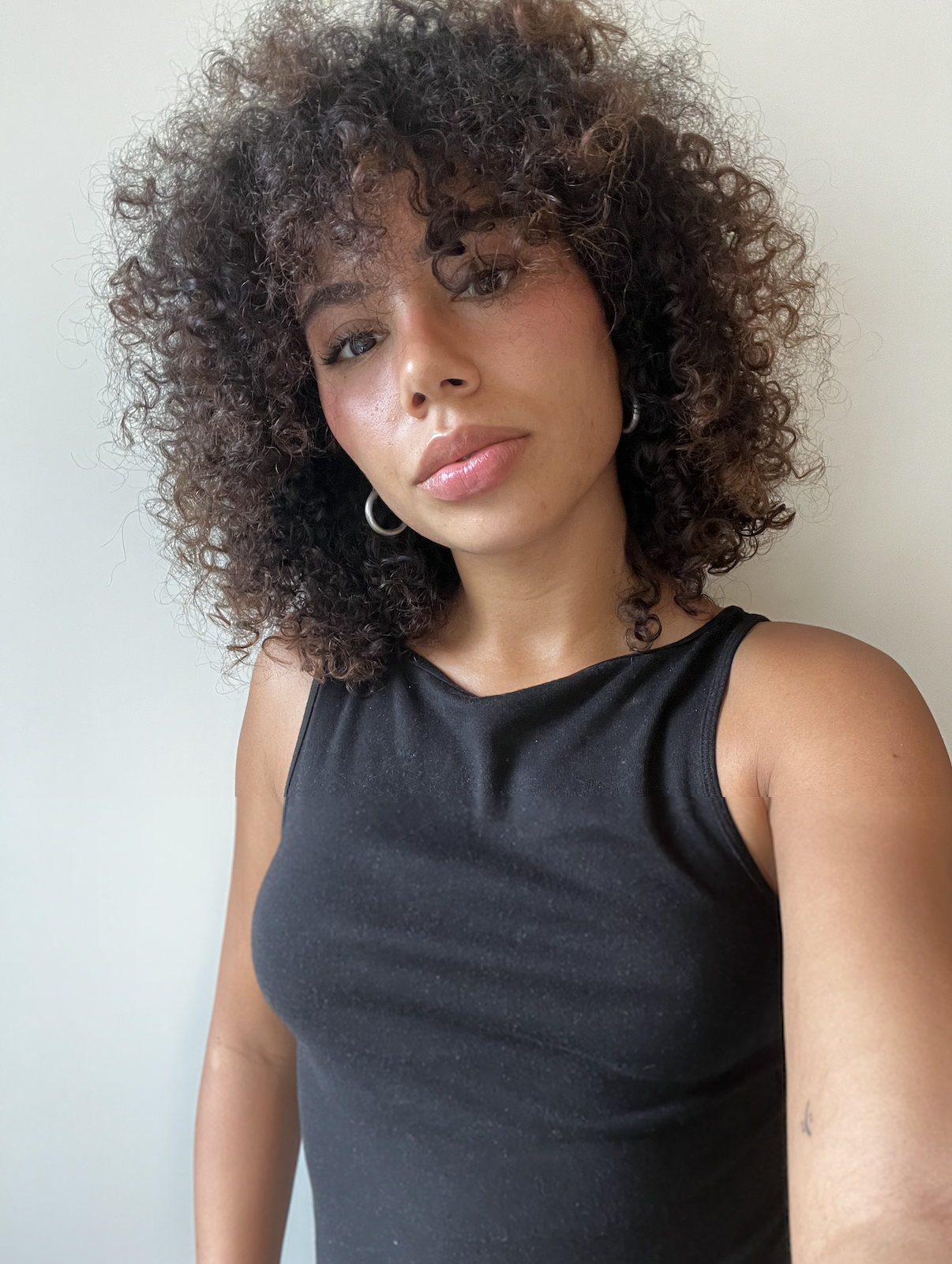 A woman with curly hair wearing a black sleeveless top and hoop earrings, standing against a plain light-colored wall.