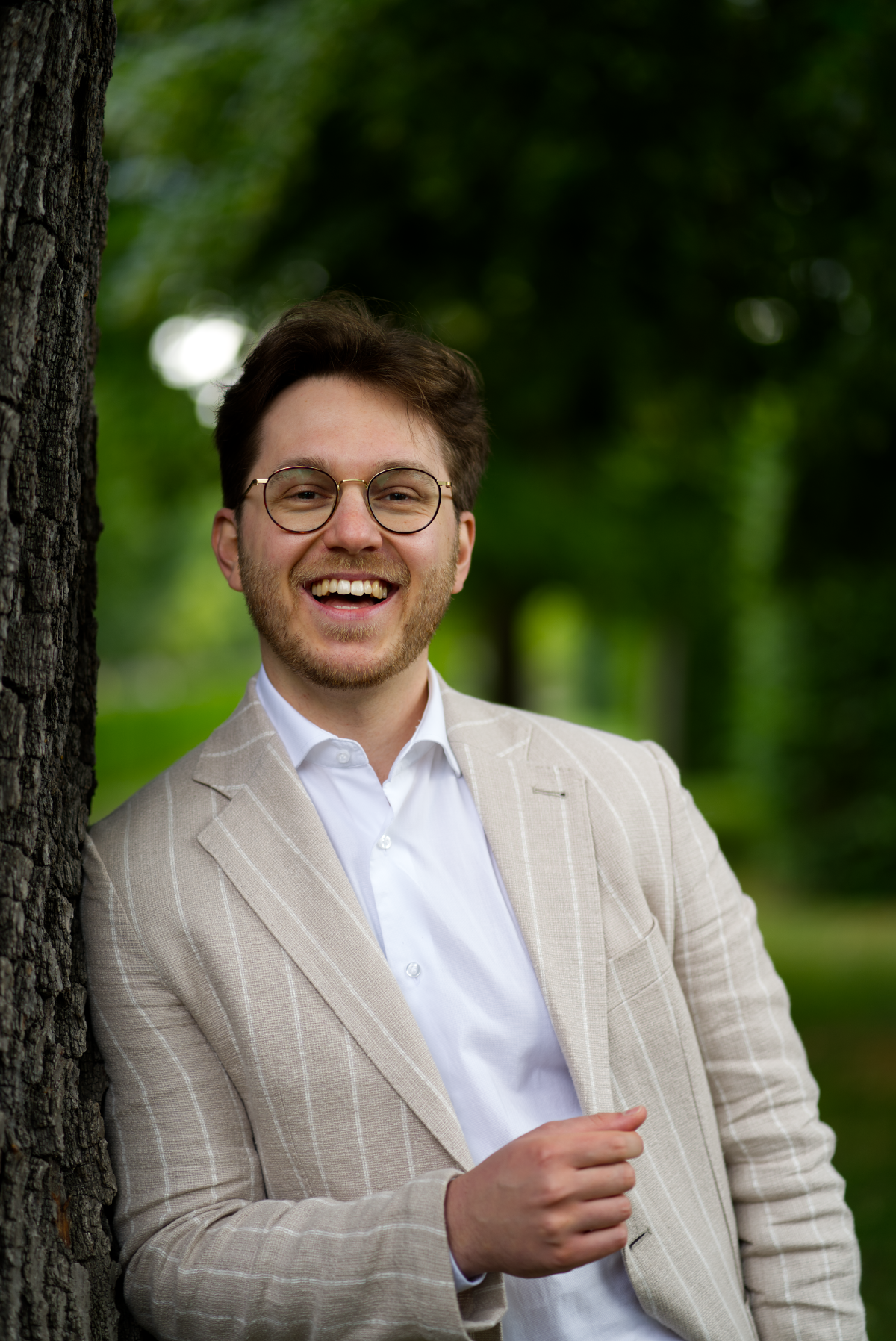 Freier Redner Alexander Bienek lächelt und lehnt an einem Baum. Er trägt ein weißes Hemd und ein beiges Sakko.
